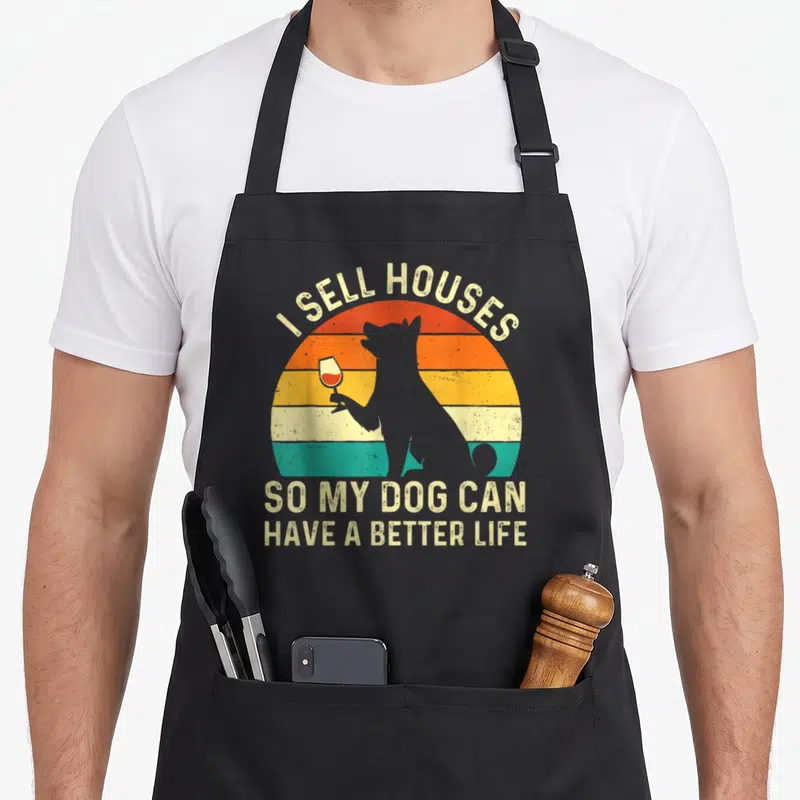 Estate dog apron - Grey dog silhouette holding wine glass against striped sunset with text I sell houses so my dog can — shown on a black cotton canvas apron against a white background
