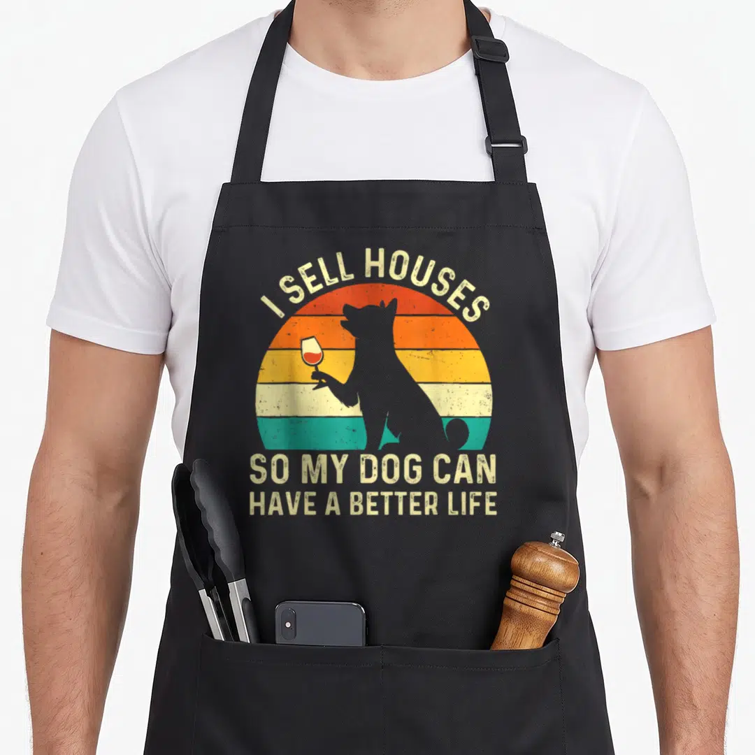 Estate dog apron - Grey dog silhouette holding wine glass against striped sunset with text I sell houses so my dog can — shown on a black cotton canvas apron against a white background