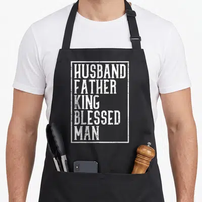 Husband king apron - White distressed stacked text HUSBAND FATHER KING BLESSED MAN inside a rectangular border on black — shown on a black cotton canvas apron against a white background