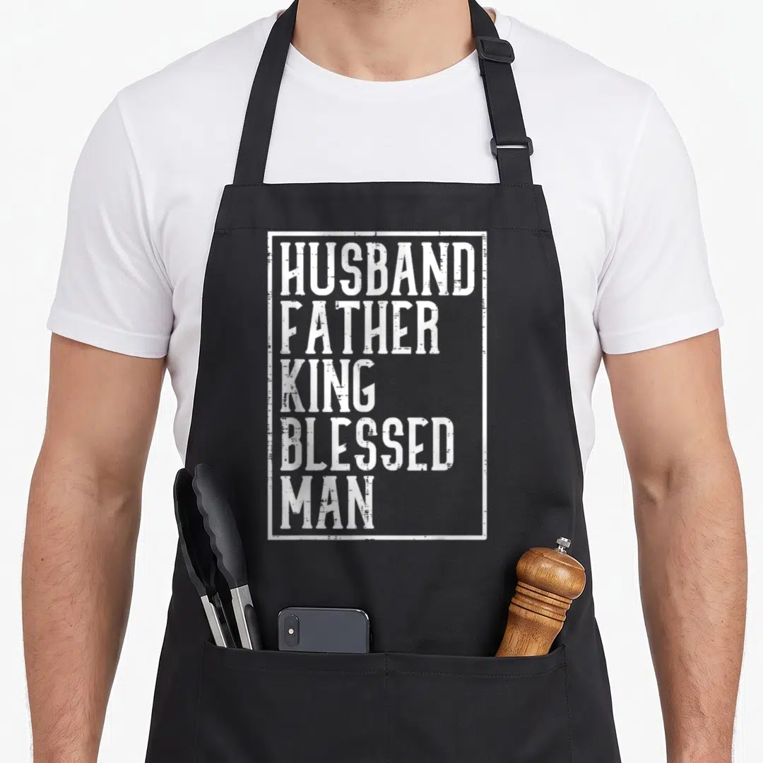 Husband king apron - White distressed stacked text HUSBAND FATHER KING BLESSED MAN inside a rectangular border on black — shown on a black cotton canvas apron against a white background