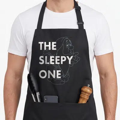 Outlined snow white apron - White THE SLEEPY ONE text over grey line art of a pointing sleepy character — shown on a black cotton canvas apron against a white background