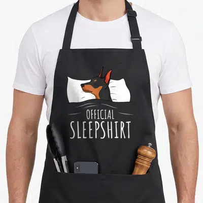 Min dog apron - Cartoon black and tan dog sleeping on white pillow under grey blanket with text OFFICIAL SLEEPSHIRT — shown on a black cotton canvas apron against a white background