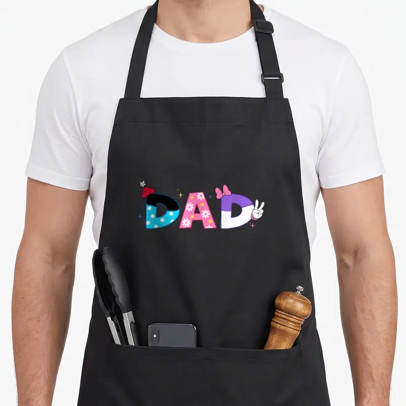 Cool birthday father day apron - Cartoon DAD text with hat, flower, bow, peace sign hand in blue pink purple red — shown on a black cotton canvas apron against a white background