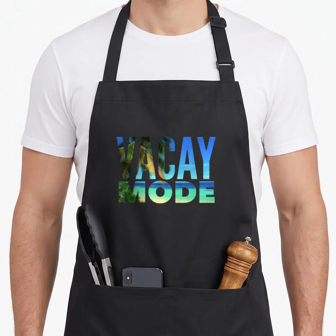Mode tree apron - VACAY MODE text filled with blue sky green palms and yellow sand beach scene — shown on a black cotton canvas apron against a white background