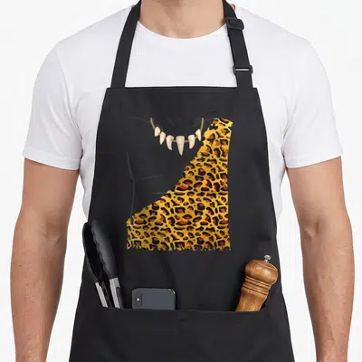 Caveman halloween apron - Cartoon torso with tooth necklace and leopard skin cloak on grey background — shown on a black cotton canvas apron against a white background