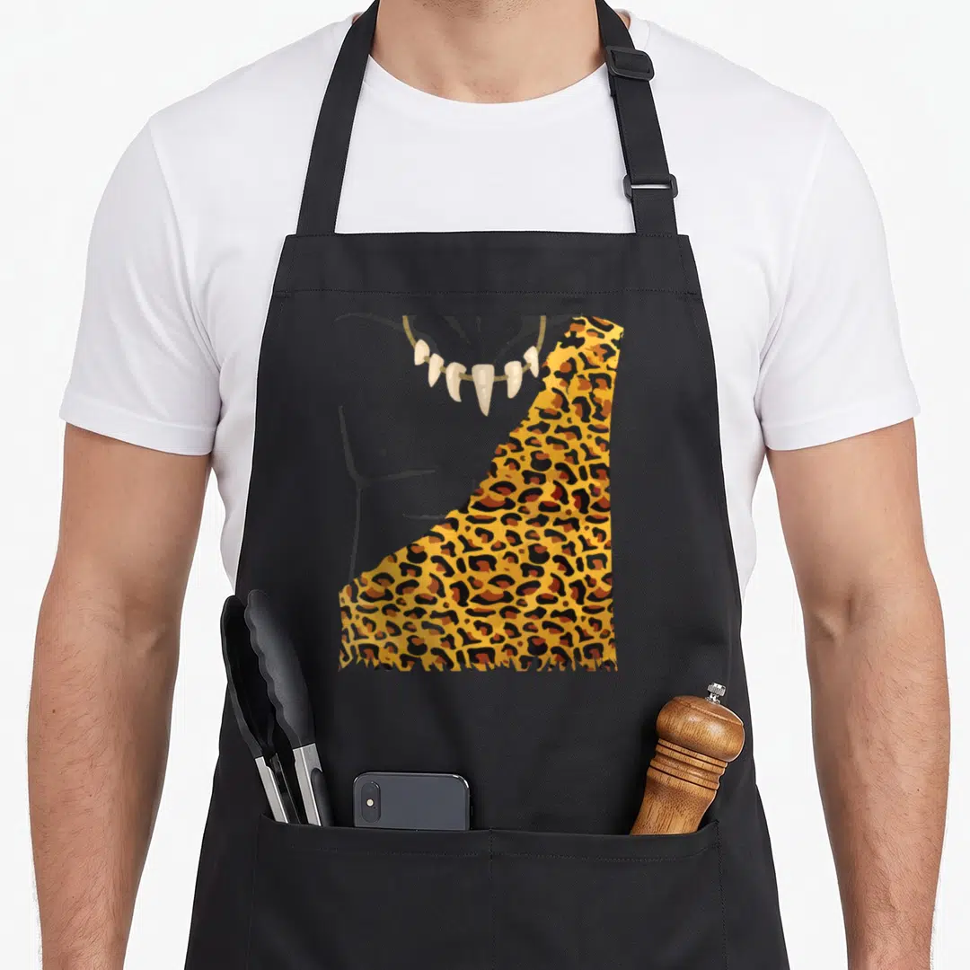 Caveman halloween apron - Cartoon torso with tooth necklace and leopard skin cloak on grey background — shown on a black cotton canvas apron against a white background