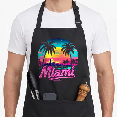 Miami beach florida apron - Retro pink and yellow Miami Florida sunset with palm trees and skyline silhouette — shown on a black cotton canvas apron against a white background