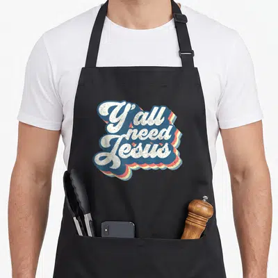 70s christian apron - Y'all need Jesus white text with blue red yellow retro shadow — shown on a black cotton canvas apron against a white background
