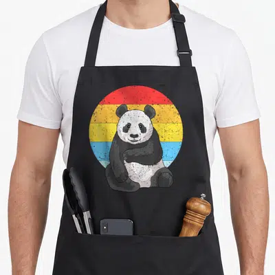 Sunset kawaii apron - Retro panda sitting before a striped circle in red yellow and blue — shown on a black cotton canvas apron against a white background