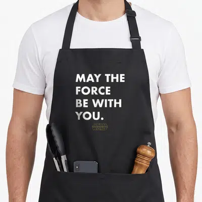 Princess essential apron - White stacked text reading MAY THE FORCE BE WITH YOU above a yellow STAR WARS logo outline — shown on a black cotton canvas apron against a white background