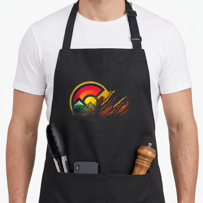 Amphitheater state colorado apron - Circular abstract logo with mountain peak and jagged brushstrokes in red, yellow — shown on a black cotton canvas apron against a white background