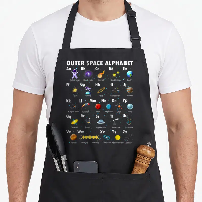 Outer kindergarten teacher apron - Outer space alphabet grid with cartoon astronauts planets stars and spacecraft — shown on a black cotton canvas apron against a white background