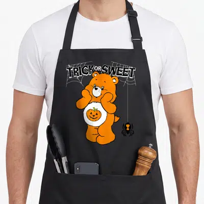 Trick apron or apron - Orange Care Bear with pumpkin belly under Trick or Sweet text and hanging spider — shown on a black cotton canvas apron against a white background