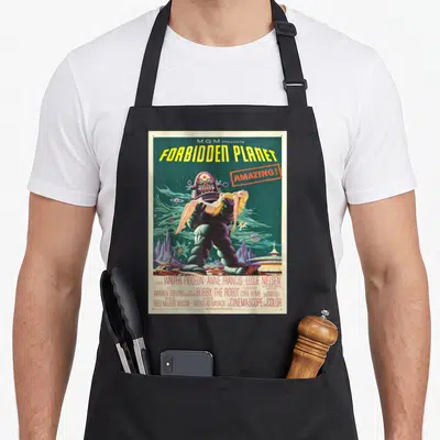 Forbidden cartoon apron - Robot carrying woman on Forbidden Planet poster with cast credits in retro colors — shown on a black cotton canvas apron against a white background