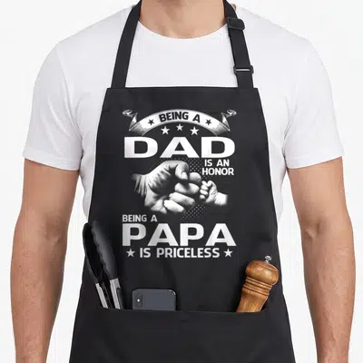 Priceless papa apron - White text and fist bump photo reading Being a Dad is an Honor Being a Papa is Priceless — shown on a black cotton canvas apron against a white background
