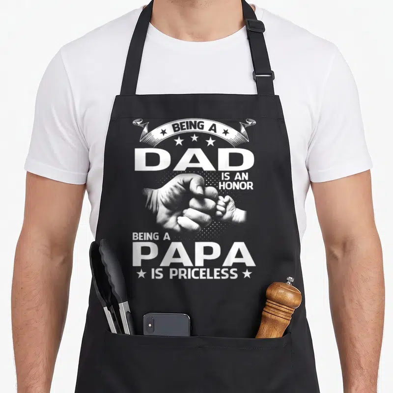 Priceless papa apron - White text and fist bump photo reading Being a Dad is an Honor Being a Papa is Priceless — shown on a black cotton canvas apron against a white background