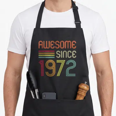 52nd birthday apron - Stacked retro typography reading AWESOME SINCE 1972 with multi-colored horizontal stripes — shown on a black cotton canvas apron against a white background