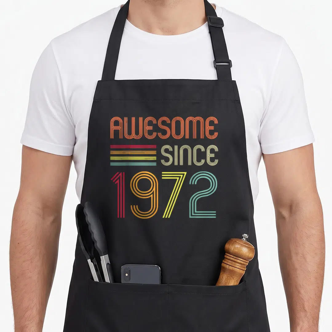 52nd birthday apron - Stacked retro typography reading AWESOME SINCE 1972 with multi-colored horizontal stripes — shown on a black cotton canvas apron against a white background