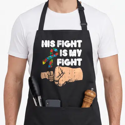 Fist ribbon apron - His Fight Is My Fight text with puzzle ribbon and peach fist bump graphic — shown on a black cotton canvas apron against a white background