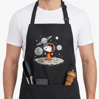 Starfield planet apron - Snoopy in orange spacesuit on grey moon with striped planets and stars — shown on a black cotton canvas apron against a white background