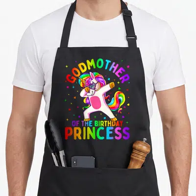 Godmother princess apron - Dabbing unicorn with rainbow GODMOTHER OF THE BIRTHDAY PRINCESS text and scattered hearts — shown on a black cotton canvas apron against a white background