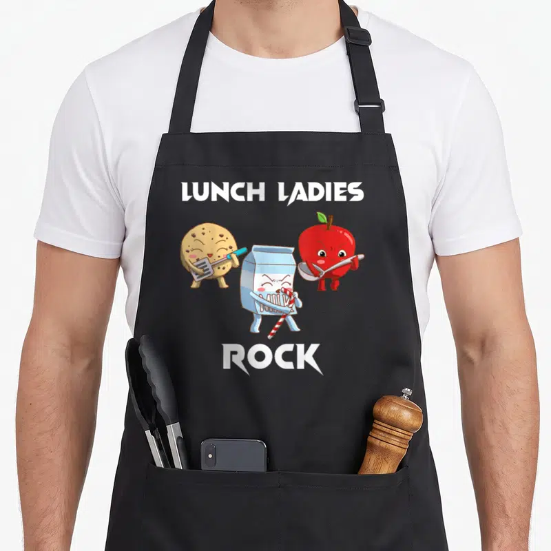 Funny rock lunch lady apron - Three cartoon food characters holding utensils under LUNCH LADIES ROCK text — shown on a black cotton canvas apron against a white background