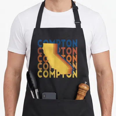 Repeat compton apron - Five stacked COMPTON words in blue red orange yellow with yellow California silhouette — shown on a black cotton canvas apron against a white background