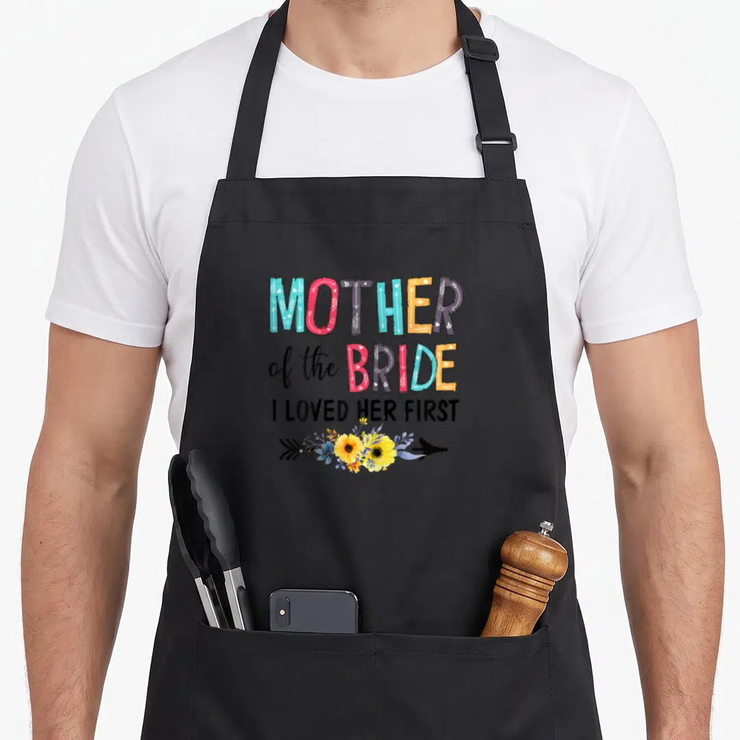 Vm6k wedding day aprons - Stacked colorful text reading Mother of the Bride I Loved Her First with floral arrow — shown on a black cotton canvas apron against a white background