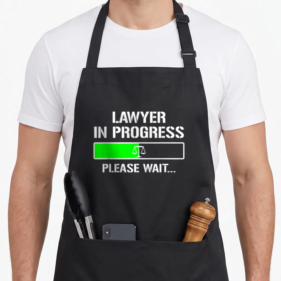Progress lawyer apron - White text reading LAWYER IN PROGRESS above a green loading bar with a scales of justice icon — shown on a black cotton canvas apron against a white background