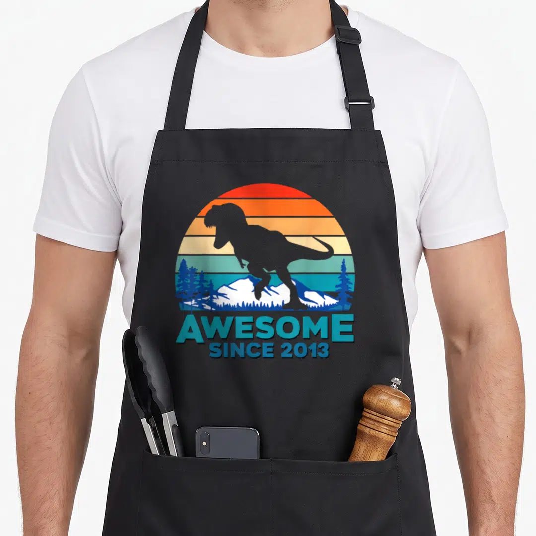 Funny dinosaur apron - Grey T-Rex silhouette over striped sun and mountains with AWESOME SINCE 2013 text — shown on a black cotton canvas apron against a white background