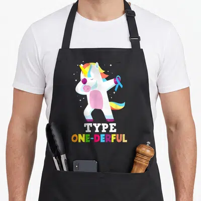 Diabetic unicorn apron - Cartoon unicorn holding blue ribbon above stacked TYPE ONE-DERFUL text in rainbow and white — shown on a black cotton canvas apron against a white background