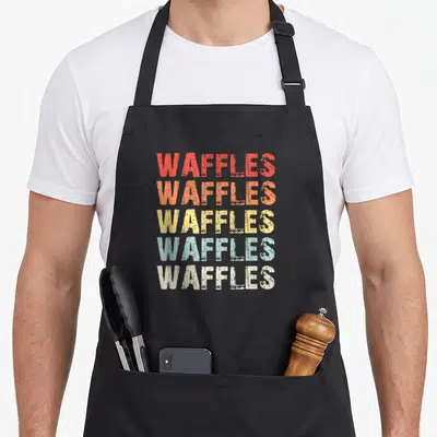 Vintage waffles apron - Stacked WAFFLES text in red orange yellow teal and beige distressed font — shown on a black cotton canvas apron against a white background