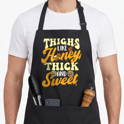 Honey thick aprons - Stacked orange and yellow text reading Thighs Like Honey Thick And Sweet with bee and daisies — shown on a black cotton canvas apron against a white background
