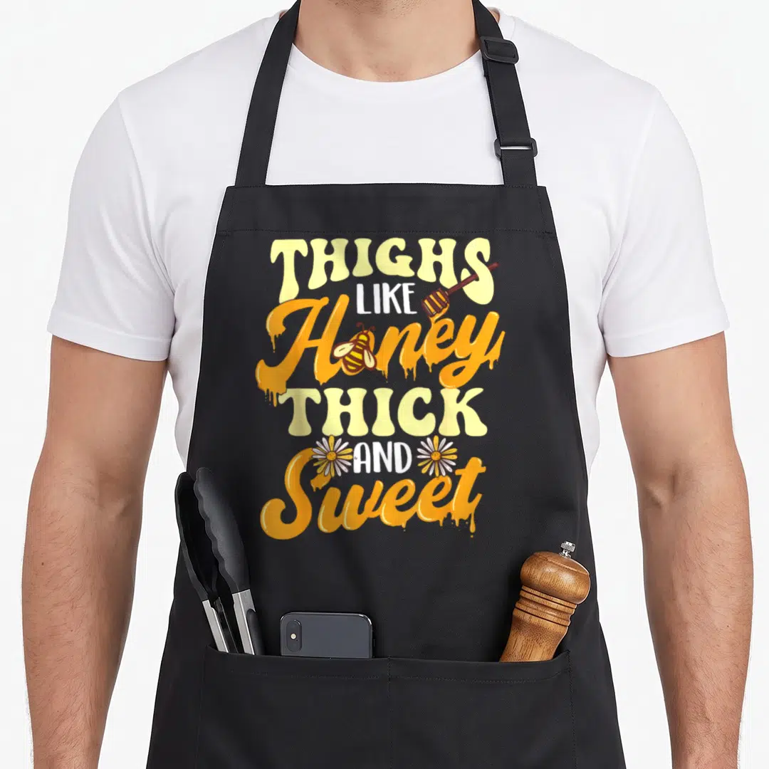 Honey thick aprons - Stacked orange and yellow text reading Thighs Like Honey Thick And Sweet with bee and daisies — shown on a black cotton canvas apron against a white background