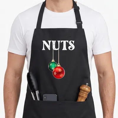 Retro chest couples christmas aprons - White NUTS text with red and green Christmas ornaments hanging by gold strings — shown on a black cotton canvas apron against a white background