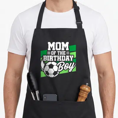 Vintage soccer birthday apron - Stacked white text MOM OF THE BIRTHDAY Boy with soccer ball and green stripes — shown on a black cotton canvas apron against a white background