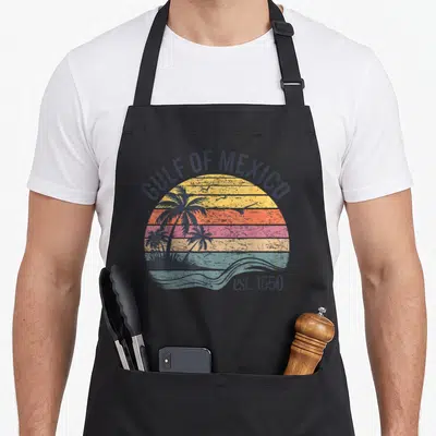 Cute gulf mexico apron - Retro sunset with palm trees, flying birds, and GULF OF MEXICO text in dark blue, yellow — shown on a black cotton canvas apron against a white background