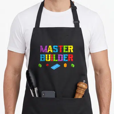Builder block grill master apron - Stacked MASTER BUILDER text made of purple yellow blue red green and orange toy — shown on a black cotton canvas apron against a white background