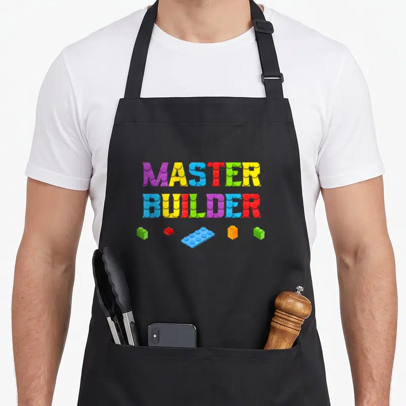 Builder block grill master apron - Stacked MASTER BUILDER text made of purple yellow blue red green and orange toy — shown on a black cotton canvas apron against a white background