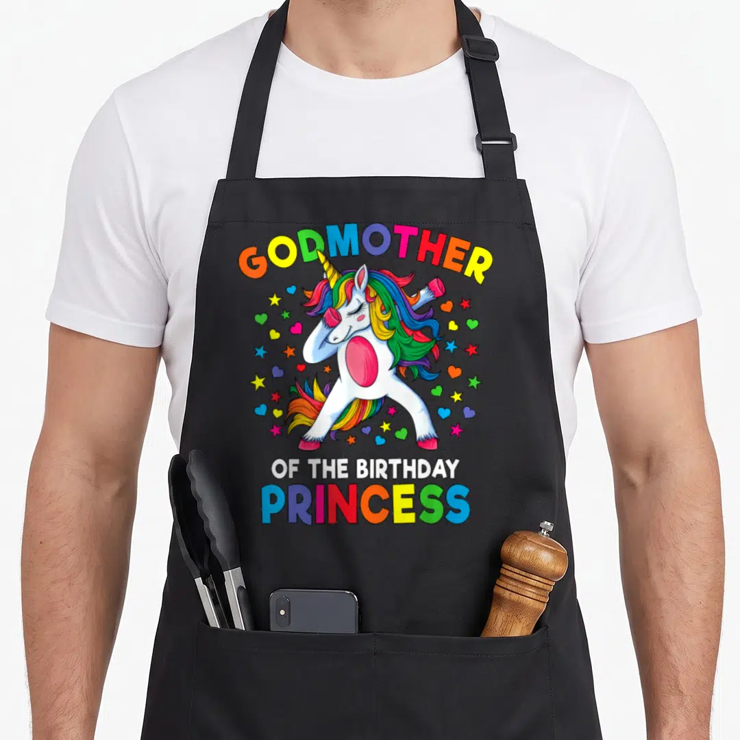 Godmother birthday princess apron - Dabbing unicorn with rainbow hair and scattered stars under arched GODMOTHER text — shown on a black cotton canvas apron against a white background