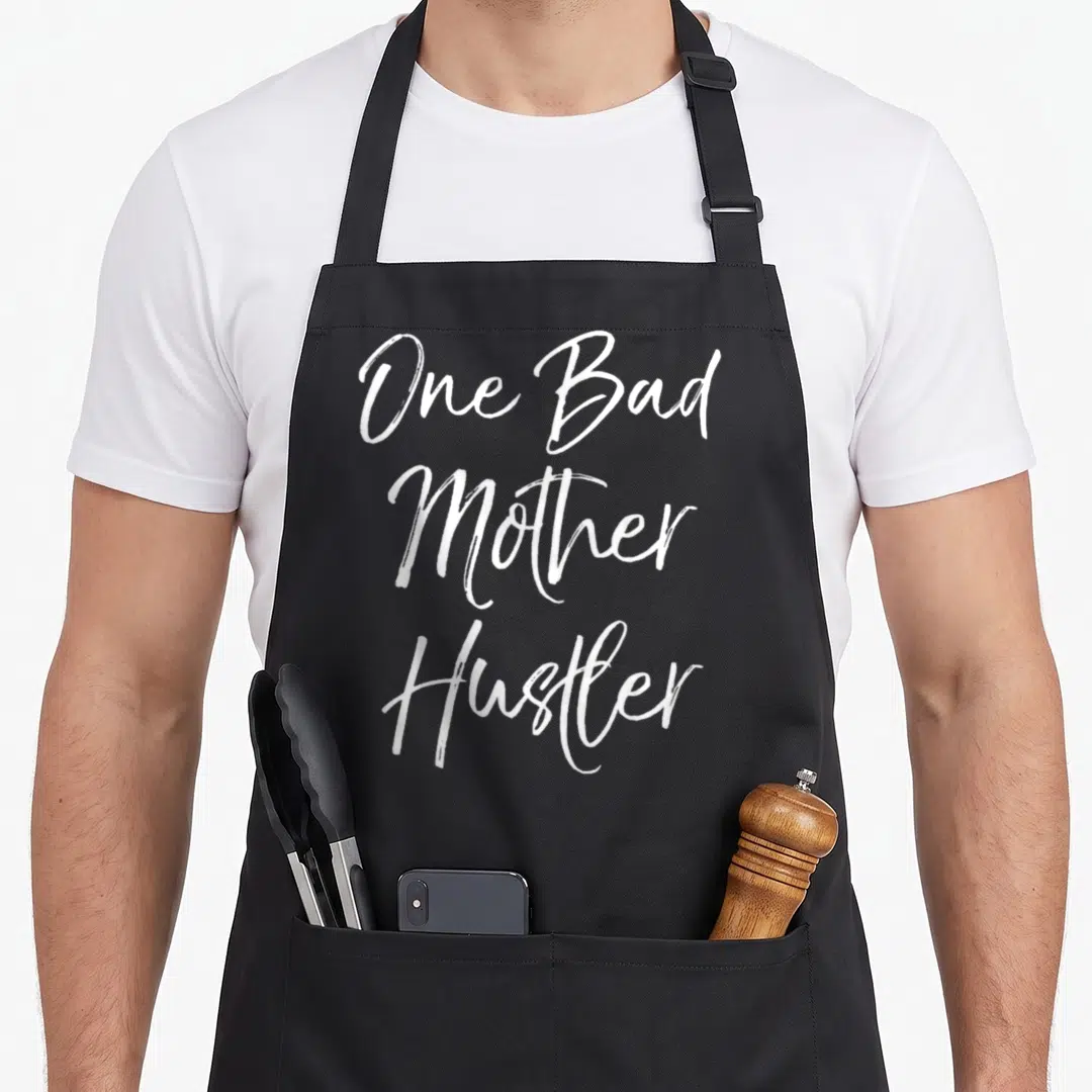 Funny hustler mother's apron - White calligraphy text One Bad Mother Hustler stacked vertically on black background — shown on a black cotton canvas apron against a white background