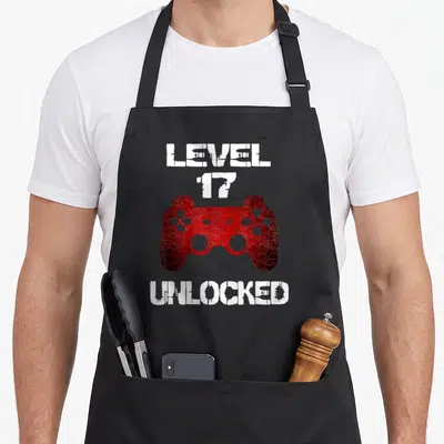 Unlocked gamer birthday apron - Red distressed game controller flanked by white LEVEL 17 and UNLOCKED text — shown on a black cotton canvas apron against a white background