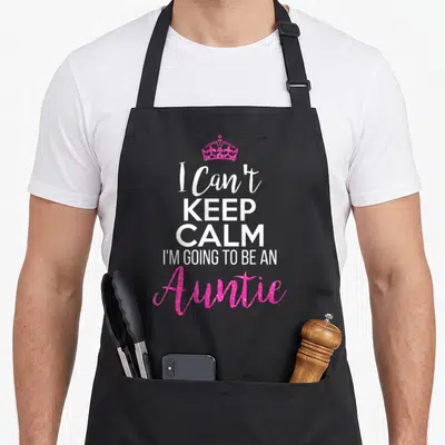 Funny auntie apron - Pink glittery crown above white and pink text reading I Can't Keep Calm I'm Going to Be an Auntie — shown on a black cotton canvas apron against a white background