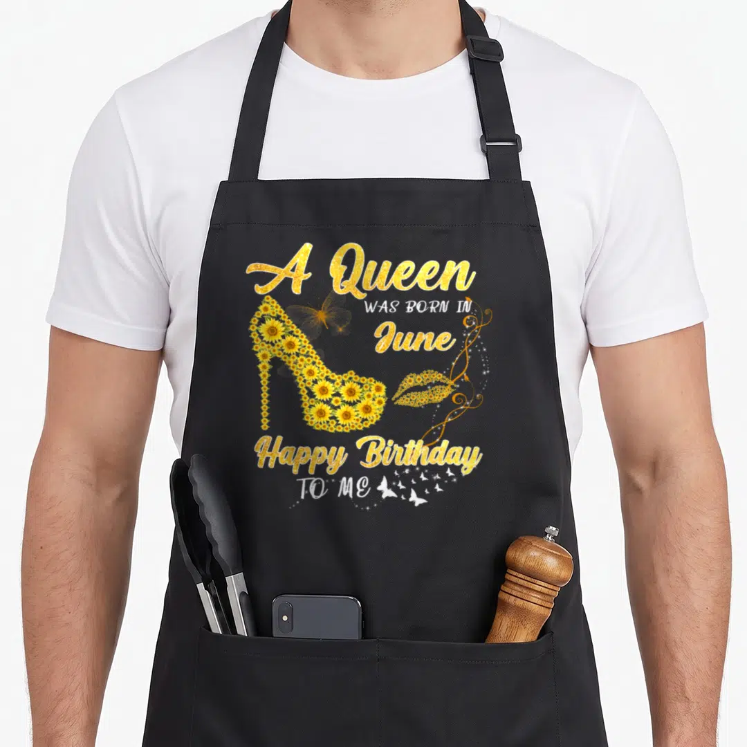 June baking queen apron - Gold sunflower high heel shoe with A Queen WAS BORN IN June text and sparkles — shown on a black cotton canvas apron against a white background