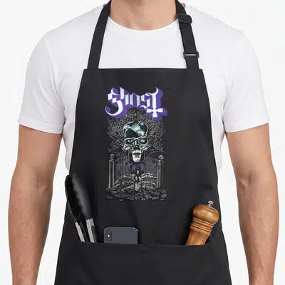 Gate ghost apron - Arched purple and white text above a winged skull behind a cemetery fence with skeletal hands — shown on a black cotton canvas apron against a white background