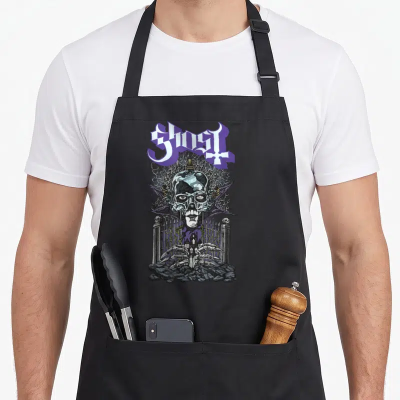 Gate ghost apron - Arched purple and white text above a winged skull behind a cemetery fence with skeletal hands — shown on a black cotton canvas apron against a white background