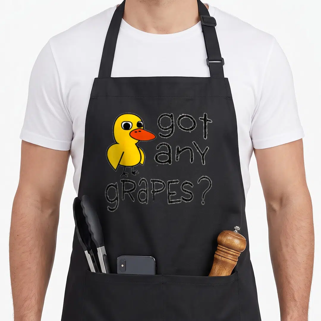 Grape duck apron - Cartoon yellow duck with orange beak next to stacked black text reading got any grapes — shown on a black cotton canvas apron against a white background