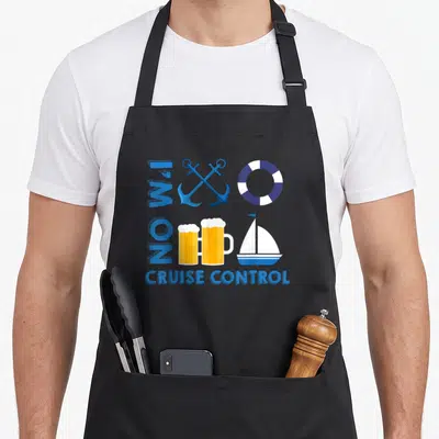 Cruise trip vacation apron - I'M ON CRUISE CONTROL text with anchors, lifebuoy, beer mugs, and sailboat in blue white — shown on a black cotton canvas apron against a white background