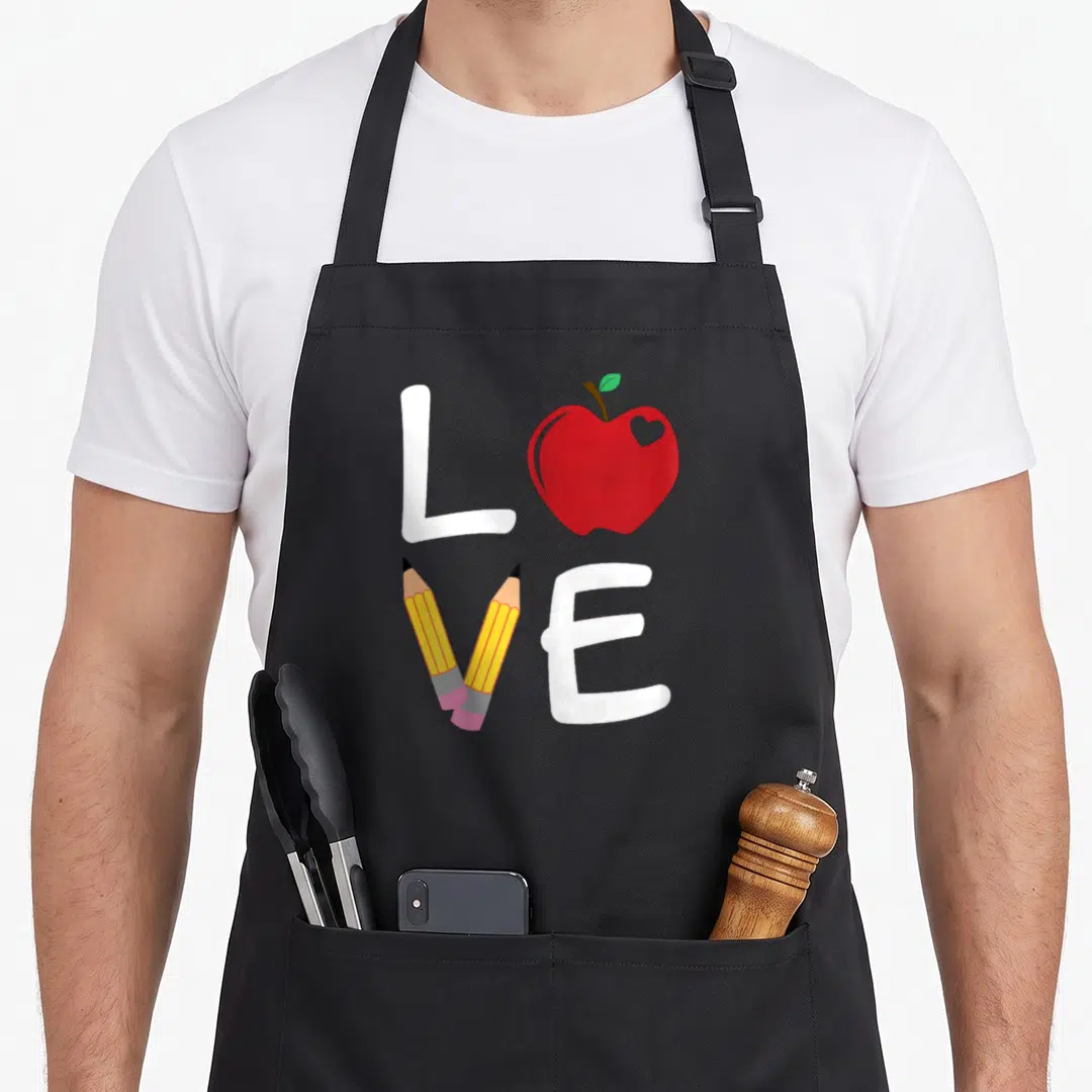 Teaching apple apron - Cartoon LOVE text with red apple replacing O and two yellow pencils forming V — shown on a black cotton canvas apron against a white background