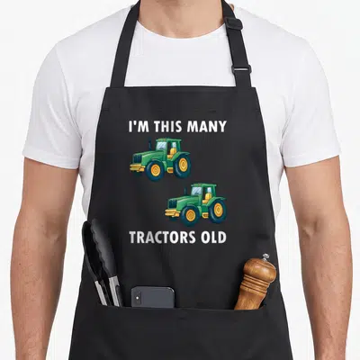 Tractor 2nd apron - Two stacked green tractors with white text I'M THIS MANY TRACTORS OLD — shown on a black cotton canvas apron against a white background